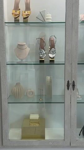 The image displays a close-up of a distressed white display cabinet with glass shelves, showcasing an array of luxury accessories and decorative objects, likely within a boutique setting in Beverly Hills.

On the top shelf, two pairs of elegant gold high-heeled sandals with pointed toes and slender straps are presented on gold risers. Beside them, a small white geometric sculpture and a ridged white candle add a touch of modern decor.

The second shelf features a pair of eye-catching silver high-heeled sandals, each adorned with a prominent flower or rose embellishment on the front strap, also displayed on gold risers. To the left, a light beige jewelry bust showcases a simple gold chain necklace.

The third shelf contains several decorative items: a light beige fluted vase, a matching ring-shaped object, a tall gold jewelry stand, and a white rectangular jewelry display stand holding delicate necklaces.

The bottom shelf features a large, rectangular gold box, upon which rests a rolled white item (possibly a jewelry cloth) and a delicate gold bracelet.

The cabinet itself exhibits a distinctive crackled or aged white paint finish, giving it a vintage or shabby-chic appearance. Its doors are fitted with dark, ornate metal handles. The items are neatly arranged against a plain white background inside the cabinet. There is no visible text in the image.