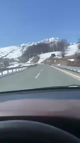 The video shows a car driving on an asphalt road through a mountainous area. Patches of snow cover the surrounding hillsides and mountains, and a small wooden structure is visible on a nearby slope. The sky is clear blue. The video is shot from inside the car, capturing the road ahead through the windshield. A few trees stand near the road, some bare, some with foliage. The car moves steadily along the road, which has a white dashed line dividing the lanes and a metal guardrail on the side.