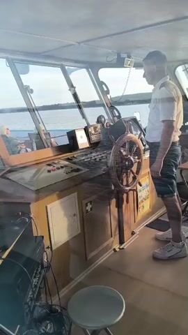 A person wearing a white shirt and dark shorts stands on a boat's bridge, holding a wooden steering wheel. The bridge is equipped with various control panels and instruments. The person appears to be steering the boat, with the water and shoreline visible through the windows.