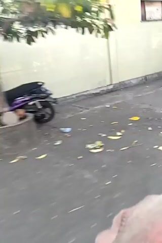 The video shows two men on motorcycles driving down a street. The first man is wearing a blue shirt and a dark baseball cap. He is riding a white motorcycle. The second man is wearing a black t-shirt with yellow writing on the back and a grey baseball cap. He is riding a black motorcycle. The street is paved with asphalt and has a concrete sidewalk. There are buildings on either side of the street, some of which appear to be shops or businesses. The buildings are painted in various colors, including light green, yellow, and orange. There is also some construction scaffolding visible in one of the buildings. The video is shot from the perspective of someone on a motorcycle, looking forward.