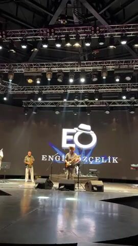 The video shows a live music performance on a stage. In the background, a large screen displays a logo with the letters "EO" and the name "ENGİNOZCELİK" below it. The stage is illuminated by numerous overhead lights and red stage lighting. There are four musicians on stage. On the far left, a person is playing a stringed instrument, possibly a guitar. Next to them, a person is playing a saxophone. In the center, a person is playing a guitar and singing. To the right of the singer, another person is playing a musical instrument, possibly a keyboard or another stringed instrument, and there are two speakers and a music stand on the stage. The overall scene suggests a professional music event.