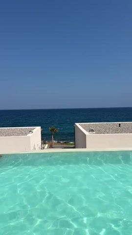 The video shows a clear blue sky above a vast expanse of deep blue ocean. In the foreground, a bright turquoise swimming pool reflects the sunlight. Two white buildings with stone accents flank the view of the ocean. A lone palm tree stands on a rocky shore between the buildings, with the ocean waves gently lapping at the coastline. A yellow buoy is visible in the water.