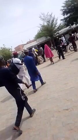 The video shows a street scene with several people walking and riding motorcycles. Many are wearing traditional clothing. The area appears to be a town square or public gathering space with buildings and trees in the background. The camera follows people walking. Later, it shows a man on a motorcycle entering an archway.