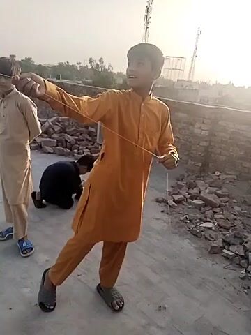 The video shows three young boys on a rooftop, with one boy in the foreground dressed in an orange shalwar kameez holding a string, possibly for flying a kite. He is looking towards the right side of the frame. To the left of him stands another boy in a beige shalwar kameez, looking in the same direction. Behind the boy in orange, a third boy is kneeling on the ground, possibly adjusting something. The rooftop is made of concrete and is littered with bricks and debris. In the background, there are buildings, trees, and antennas visible against a bright sky. The sun is shining, creating a hazy atmosphere.