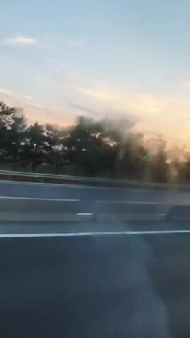 The video shows a view from inside a vehicle looking out at a highway during sunset. The sky is a mix of blue and orange, with some clouds. Trees are visible along the side of the highway, and there are concrete barriers separating the lanes. A few cars are driving on the highway in the distance. The image is slightly blurry, suggesting the vehicle is in motion.