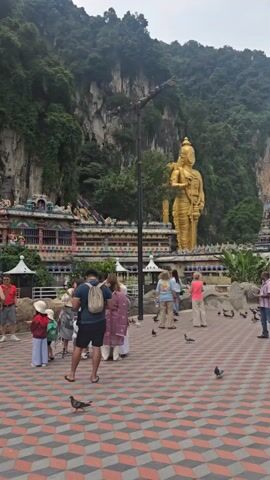The video shows a wide shot of a Hindu temple complex in front of a massive limestone hill. Several people are standing and walking on a geometrically-patterned brick plaza. Many pigeons are walking around the plaza. A towering golden statue of a Hindu deity stands prominently in the background. The sky is overcast.