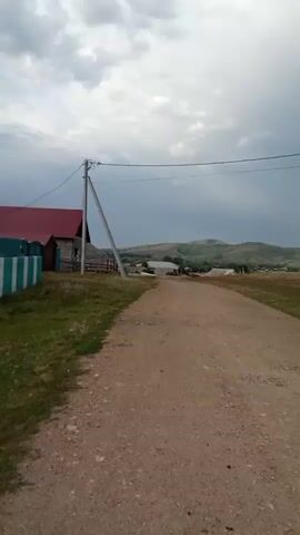 A dirt road leads into a rural landscape under a cloudy sky. On the left side of the road, there is a wooden building with a red roof, a fence, and a utility pole. The road continues into the distance, passing through a field with some scattered buildings. In the background, rolling hills are visible, and a faint rainbow arcs across the sky. The overall impression is one of a serene, rural setting, possibly just after a rain shower.