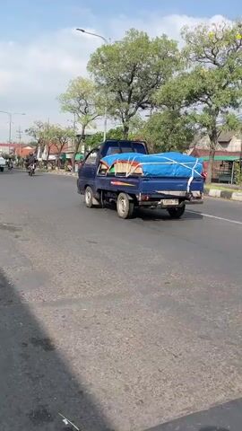 A blue pickup truck is driving on a paved road under a clear sky. The truck bed is loaded with items covered by a blue tarp, secured with rope. The truck is in the right lane of traffic. A black car is partially visible in the right lane, ahead and to the right of the pickup truck. To the left of the road, there are trees, buildings, and utility poles. The road appears to be in a semi-urban or suburban area.