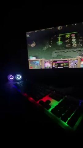 In a darkened room, a computer monitor displays the real-time strategy game Warcraft III. The game screen shows a top-down perspective of a map with various buildings and units.  The interface at the bottom displays player controls and resources.  Below the monitor is a gaming keyboard illuminated with red, green, and blue LEDs.  A mouse is also visible to the right of the keyboard.  The overall scene suggests someone is playing or about to play the game.