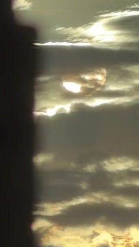 This is a vertical, close-up, time-lapse video of the sun partially obscured by clouds. The sun appears as a bright, glowing disc with patchy clouds drifting across its surface. The clouds themselves are dark and dense in the foreground, with lighter, wispy clouds in the background. The overall color palette is muted, with a mix of grays, whites, and yellow hues. The video captures the subtle movement of the clouds, which gradually shift and change the appearance of the sun. The perspective is fixed, with no apparent camera movement.