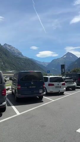 The video shows a parking lot with several cars. In the background, there are mountains and a clear blue sky with some clouds. There is a building with the "SOCAR" logo on it. Some people are walking towards the building. The scene appears to be in a mountainous region during the daytime.