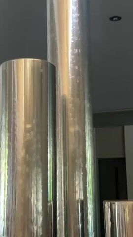 The video shows a close-up view of several large, cylindrical objects covered in a reflective, metallic material. The objects appear to be arranged vertically. The surface of the material has a textured, crinkled appearance, suggesting it might be foil or a similar wrapping. Light reflects off the surfaces, creating bright highlights and shadows, which obscure some details. In the background, a dark doorway or opening is visible, as well as what appears to be the ceiling with a light fixture. The bottom of the cylinders rests on a surface that appears to be covered in small stones or gravel. The overall impression is of a display or installation featuring these metallic cylinders. There are no people or animals visible in the video.