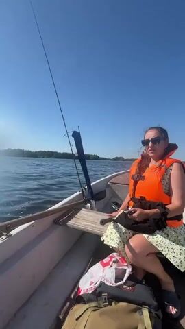 The video begins with a view from inside a boat, showing a woman wearing an orange life vest and sunglasses. She is sitting on a bench and looking to her right. A fishing rod is visible in the boat. The boat is on a body of water with a clear blue sky above. The scene then shifts to a view from the back of the boat, showing the water churning as the boat moves forward. The sun is visible in the sky, and the horizon shows a line of trees. The boat continues to move through the water, creating a wake. The video captures the experience of being on a boat on a sunny day.