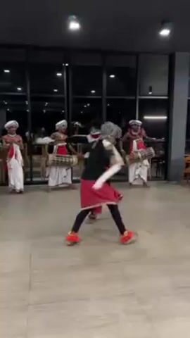 The video shows a traditional Sri Lankan dance performance. Two dancers in elaborate costumes are at the center of the frame, executing swift, synchronized movements. They wear red skirts, black tops, and distinctive masks with white embellishments resembling birds. They perform barefoot on a light-colored tiled floor. Behind them, four musicians play drums, also dressed in white traditional clothing. The environment appears to be a modern building with large windows offering a glimpse of the outdoors. The lighting is bright and even.