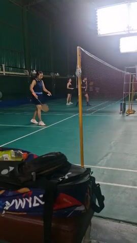 The video shows a badminton court with several people playing. In the foreground, there are badminton bags on a bench. In the background, people are actively engaged in a badminton game, hitting the shuttlecock back and forth over the net. The court has green flooring and green walls. There are badminton nets set up across the court. The lighting appears to be artificial.