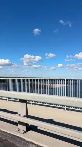 The video shows a view from a bridge looking out over a body of water on a clear day with blue skies and scattered white clouds. A metal railing runs along the edge of the bridge, with a lamppost visible in the center of the frame. The water is calm and appears to have a sandbar or shallow area visible in the distance.