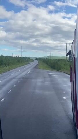 A video of a road from a vehicle's perspective. The road is wet and reflects the sky. Trees line the side of the road. The sky is blue with white clouds. A side mirror shows a reflection of the road and the vehicle.