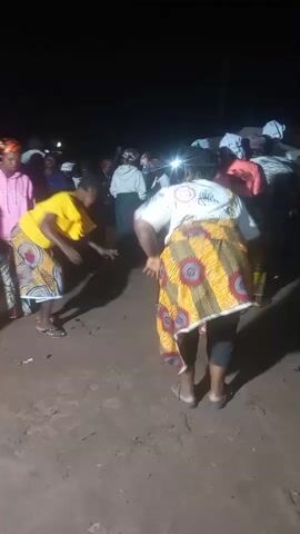 A group of women are dancing in what appears to be an outdoor nighttime setting. The ground is dirt. The lighting is dim with some artificial light sources, likely from a camera or small portable lights. The women are wearing colorful clothing, including headscarves and patterned skirts. One woman in the foreground is wearing a yellow shirt and a patterned skirt. Another woman to her right is wearing a white shirt with a patterned skirt and is bent over. Behind them, other women are also dancing and observing. The overall atmosphere seems to be festive.