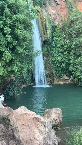 A video shows a natural waterfall cascading into a clear green pool of water. Lush green foliage and red rock formations surround the scene. Two individuals are visible near the water's edge. One person stands partially submerged in the water, facing away from the camera, with their arms crossed. Another person is seen from behind, kneeling near the water in the foreground. The camera pans slightly, capturing the expansive greenery and the raw beauty of the natural landscape.