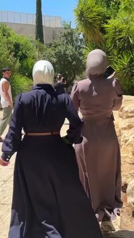 Two women in hijabs are walking away from the camera down a cobblestone path. The woman on the left wears a dark navy dress with a brown belt and a white hijab. The woman on the right wears a mauve dress with a brown belt and a mauve hijab. Both are wearing sneakers. In the background, there are trees, some people walking, and buildings. The scene appears to be outdoors on a sunny day. The location is identified as Al-Eizariya, Palestinian Territories.