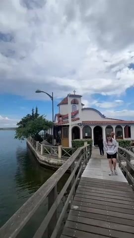 The video shows a small white and blue church with a red-tiled roof situated on a small peninsula extending into a large body of water. A wooden boardwalk with a railing extends from the mainland to the church. The sky above is partly cloudy with patches of blue sky visible. The water is relatively calm with some ripples on the surface. In the distance, the shoreline is visible. The perspective shifts to show the vastness of the water and the sky. Later in the video, the view is from the boardwalk, showing the church at the end of the pier and the surrounding lake and sky.