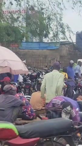 The video shows a street scene with several motorcycles parked in a cluttered area. In the background, a building with the sign "NESTANG PLACE" is visible. People are gathered around the motorcycles, some appearing to be interacting with them. To the right, there are makeshift stalls with goods displayed, including what appear to be large water containers. Further in the background, there are more buildings and a water tank. The environment appears to be a busy urban or semi-urban area with evidence of both commerce and residential structures. The video also captures reflections on a surface, possibly a window, obscuring parts of the view at times.