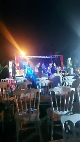 Here is a forensic description of the video:

The video captures an outdoor event at night, featuring a stage illuminated by bright lights. Several people are visible on and around the stage, but their specific actions are difficult to discern due to the distance and lighting conditions. In the foreground, numerous white chairs are arranged facing the stage, suggesting an audience seating arrangement. The chairs appear to be occupied, as people are seated in them, their backs towards the camera. The background is dark, indicating the event is taking place after sunset, with the stage lights providing the primary source of illumination. Flags and banners are placed behind the stage. The overall impression is of an organized outdoor gathering, likely a performance or presentation of some kind.