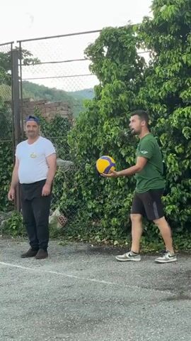 A man in a green t-shirt and dark shorts serves a blue and yellow volleyball outdoors, tossing the ball high with his left hand as he prepares to strike it with his right. He stands on a gray paved surface. To his left, another man in a white t-shirt, dark pants, and a blue baseball cap stands observing. Behind the observing man is a chain-link fence, and lush green trees and bushes fill the background.