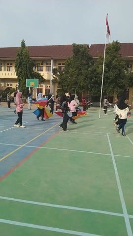 This media file captures an outdoor event featuring a group of people, likely students, participating in a flag ceremony or performance. The subjects are dressed in casual attire, with several individuals holding colorful flags that are being waved. The setting appears to be a schoolyard or athletic field, indicated by the presence of a building with a red tiled roof and basketball hoop. The ground is a green and blue court with white and colored lines, suggesting it's a sports facility. The time of day is likely morning or late afternoon, given the angle of the sunlight and the clear sky. The atmosphere seems orderly and engaged, with participants focused on their activity. One flag prominently displayed is the Indonesian flag, suggesting the event may be a national celebration or a school assembly. The overall scene depicts a structured activity, possibly a parade or a drill, where the participants are synchronized in their movements with the flags.