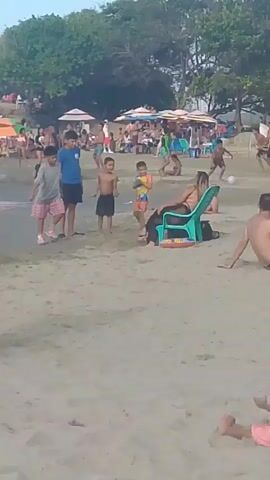 The video shows a crowded beach scene. Several children are playing in the shallow water and on the sand. Adults are scattered throughout the scene, some sitting on chairs, others standing and talking. There are colorful umbrellas set up on the beach, providing shade. The environment appears to be a tropical or sub-tropical location with lush green trees in the background. The sky is clear and blue. The overall atmosphere suggests a day of leisure and recreation.