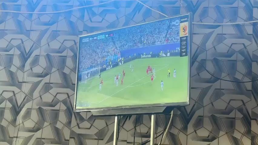 A television displays a soccer match. The game is being played on a green field with two teams wearing red and white uniforms. The score is visible in the upper left corner of the screen. The television is mounted on a black stand. The background of the image is a wall with a geometric patterned wallpaper. The lighting in the room is bright and appears to be coming from above, casting a glare on the television screen.