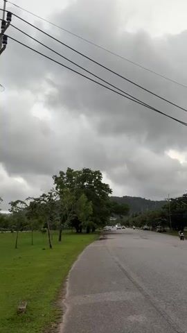 This media file captures a road scene in Ko Kaeo, Thailand, under a cloudy sky. The overcast weather suggests it might be early morning, late afternoon, or a stormy day.

The foreground shows a paved road with a yellow line dividing the lanes. The road curves to the left, bordered by a grassy area. In the middle ground, trees line the right side of the road, with some vehicles and a motorcycle visible. Further into the distance, there's a body of water, possibly a flooded area or a lake, with trees and electrical pylons in the background. An excavator is also visible near the water.

In the first frame, two people, a woman and a young girl, are seen from behind, walking towards the road. The woman has blonde hair and is wearing a black top, while the girl is wearing a pink shirt.

The overall mood is somewhat subdued due to the cloudy weather. The presence of the flooded area and the excavator suggests potential ongoing construction or the aftermath of heavy rainfall. There are no visible signs or text in the media.