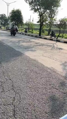 A person wearing a helmet and backpack is riding a scooter on a paved road. The road has some cracks and a shadow is cast on it. In the background, there are trees, a vehicle, and a blue object. The sun is shining, creating a bright atmosphere.