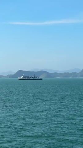 A ferry moves across a vast, blue ocean. In the background, a chain of islands with lush green mountains is visible. The sky is a clear, bright blue with a few wispy clouds. The water is a deep turquoise color and appears choppy with small waves. The ferry is white with a few dark accents and appears to be a passenger ferry. It is moving from left to right across the frame.