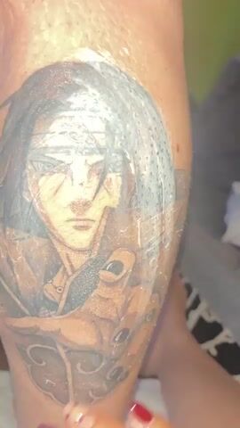 A close-up shot shows a hand with dark red fingernail polish gently rubbing a balm or lotion onto a large black and grey tattoo of a person's face and shoulders on someone's leg. The tattoo appears to be of an anime character. The hand is positioned over the upper part of the tattoo, obscuring some of the face. The background is blurred and dark, with hints of a patterned surface and possibly furniture. The lighting is dim, focused on the tattoo and the hand.