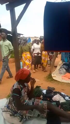 The video depicts an outdoor market scene with several people. In the foreground, a woman wearing a red headscarf and a patterned dress is sitting on the ground, surrounded by piles of clothing. She is holding something to her mouth, possibly smoking. In the background, several other women are walking and interacting in what appears to be a market stall area. The ground is dirt, and there are wooden structures overhead, suggesting a rustic marketplace. The overall atmosphere appears to be one of commerce and daily life.