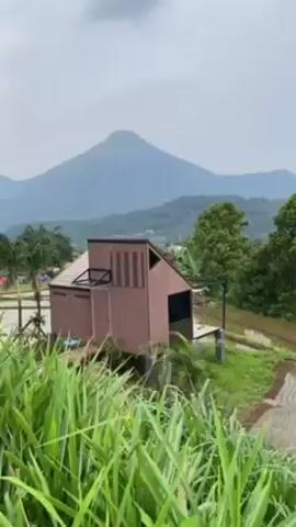 The video shows an outdoor scene with buildings and a mountain in the background.
The video starts with a close-up of a brown building with a unique architectural design. Rice fields are visible in the foreground. In the background, a mountain rises against a cloudy sky. The camera then pans to the right, showing a paved walkway with two people walking along it. The area is grassy, and there are more buildings visible. The people are walking away from the camera. The camera continues to pan, showing a wider view of the area, including more buildings on a grassy slope and more people walking around. A low wooden fence is also visible, separating different sections of the area.