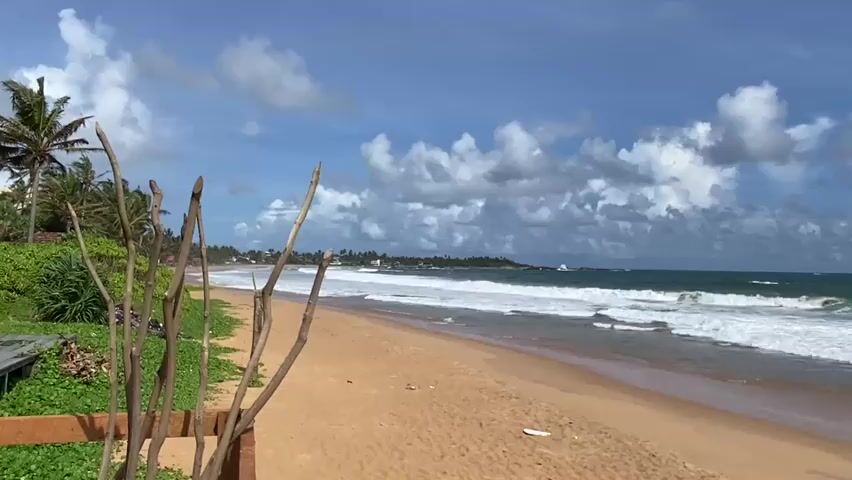 The video captures a wide shot of a sandy beach with the ocean in the background. The sky is blue with scattered white clouds. Waves are breaking on the shore, creating white foam. On the left side of the frame, there are green bushes and palm trees. In the foreground, there is a wooden fence and some dry branches. The beach appears mostly empty, with no people visible. There is a piece of white debris on the sand in the middle distance. The overall scene is a tropical beach landscape.
