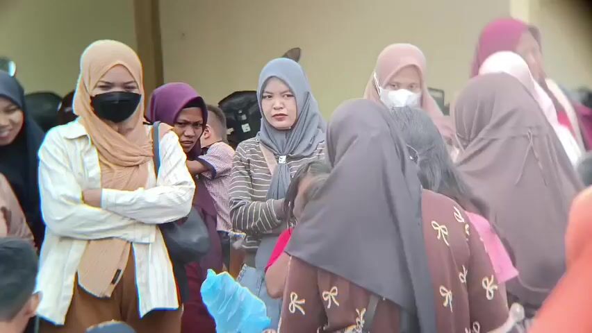 A group of people, mostly women and children, are gathered together outdoors. Many of the women are wearing hijabs. Some are looking towards the camera, while others are looking away or interacting with each other. One woman on the left is wearing a light brown hijab, a white and brown striped shirt, and a brown skirt, with her arms crossed. Next to her, a woman in a purple hijab is holding a baby. In the center, a woman in a grey hijab is looking forward. A woman in a dark brown hijab with a bow pattern on her shirt is facing away from the camera. Another woman in an orange hijab is on the far right, also facing away. The background appears to be a building with a light-colored wall. The lighting suggests it is daytime.