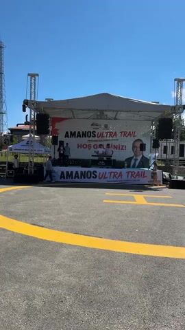 The video shows an outdoor stage set up for an event. The stage has a large banner that reads "AMANOS ULTRA TRAIL" in large, bold letters, with smaller text underneath saying "Zirvelerde Koş, Doğa'yla Buluş!" which translates to "Run on the Peaks, Meet with Nature!". There is also a portrait of a man in a suit on the banner. The stage is equipped with speakers and lighting.

Several people are visible on and around the stage. Some individuals are standing behind the banner, while others are walking in front of it. One person appears to be setting up equipment. In the foreground, there is an asphalt surface with yellow markings, suggesting it might be a parking lot or a designated area for an event. In the background, there is a tall communication tower and some buildings.

The scene is illuminated by bright sunlight, indicating it is daytime, likely late morning or afternoon, under a clear blue sky. The weather appears to be warm and sunny. The overall atmosphere seems to be one of preparation for an event, possibly a race or festival, given the "ULTRA TRAIL" and athletic imagery on the banner. The location appears to be outdoors, likely in a semi-urban or suburban area, given the presence of the tower and buildings alongside the natural-themed event branding. There are no clear signs of a crowd gathered yet, suggesting the event is either about to begin or is in its early stages.
