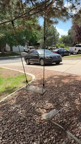 A swing hangs from a tree branch, its seat swaying gently back and forth. In the background, a dark gray sedan is parked in a driveway. Several other vehicles and houses are visible, suggesting a suburban setting. The ground is covered with gravel and some dry leaves.