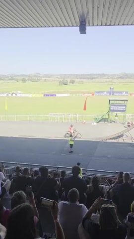 The video shows a stunt performance in an outdoor stadium. In the foreground, a crowd of spectators sits in tiered seating under a covered roof. The spectators are mostly facing away from the camera, looking towards the performance area. In the middle ground, a stunt performer on a dirt bike is in mid-air, having launched from a ramp. The performer is wearing a helmet and protective gear. Below the ramp, a car is positioned, and another performer can be seen standing near it. The background features a green field, possibly a race track or a sports field, with advertising banners visible. The sky is clear and blue, indicating a sunny day. The overall scene depicts an energetic event with an audience enjoying a motorcycle stunt show.