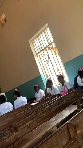 A group of people, primarily young women wearing white uniforms and red and white caps, are seated on wooden pews in what appears to be a hall or a classroom. The room has a light-colored ceiling with visible light fixtures and fans. Natural light enters through barred windows on the side of the room. The individuals are facing forward, some are looking down, possibly at notes or a book, while others are looking ahead. The focus is on the seated individuals and the pews. The camera is positioned to capture the scene from behind and to the side, with a slight upward angle. The overall impression is of a gathering or a lesson in progress.