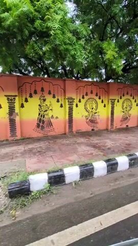 A street-level shot captures a wall adorned with colorful murals. The wall is painted with an orange and yellow gradient, and features black stencil-like artwork. The artwork depicts several figures, possibly deities or dancers, dressed in traditional attire, each with a halo around their head. Between these figures are decorative pillars and rows of bells hanging from arched borders. To the right of the mural, there's a depiction of a tree. In front of the wall, there's a sidewalk with a black and white striped curb. Beyond the curb, a wet asphalt road is visible, suggesting recent rain. Lush green trees with dense foliage overhang the wall and the scene. The camera appears to be moving, providing a panning view of the mural.