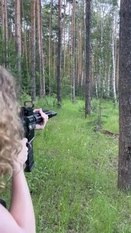 A young woman with curly blonde hair stands in a forest clearing, holding a rifle. She is wearing a gray tank top and denim shorts. She looks at the camera and smiles. The rifle is black and appears to be a semi-automatic rifle with a scope. The forest is dense with tall trees and green grass.