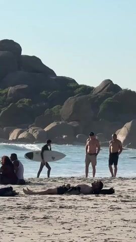 The video shows a beach on a sunny day. A man is lying down on the sand. In the background, three men are standing or walking by the water's edge. One of them is carrying a surfboard. Behind them are large rocks covered with greenery. There are also other people seated on the beach to the left of the frame. The water has small waves.
