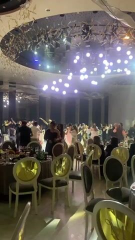 The video shows a group of people dancing at what appears to be a wedding reception.  The dancing is happening in the center of a large room with round tables and chairs arranged around the perimeter. The tables are set with dining ware.  The room has high ceilings with elaborate lighting fixtures including hanging lights and spotlights. Most of the dancers are women, wearing a variety of dresses.  Some people are seated at the tables, watching the dancing. The music is loud, creating a lively atmosphere.  The room appears to be decorated with reflective surfaces.
