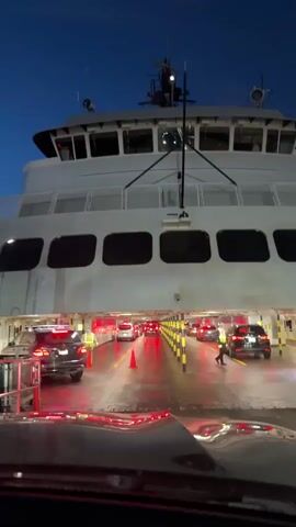 The video shows the interior of a ferry, with cars lined up and slowly moving. The camera is inside one of the cars, facing forward. There are several rows of vehicles, each guided by traffic cones and yellow lines. Ferry personnel wearing bright vests are directing the traffic. The ferry's exterior is visible, with large windows and a white hull. The sky is visible outside, suggesting it's either dawn or dusk. The ferry is on the water, with the shore visible in the background. The overall impression is of an organized and efficient ferry operation.