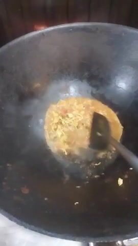 A cooking scene unfolds with a large, black wok taking center stage. Inside the wok, a bubbling mixture of noodles and what appears to be sauce is being stirred with a metal spatula. Steam rises, indicating heat, and the contents of the wok seem to be cooking. The person holding the spatula is visible only by their hand and arm. To the side of the wok, on a nearby shelf, are various containers, including bottles of soy sauce, chili sauce, and other condiments.  The background indicates the cooking is taking place in a commercial kitchen or food stall setting.