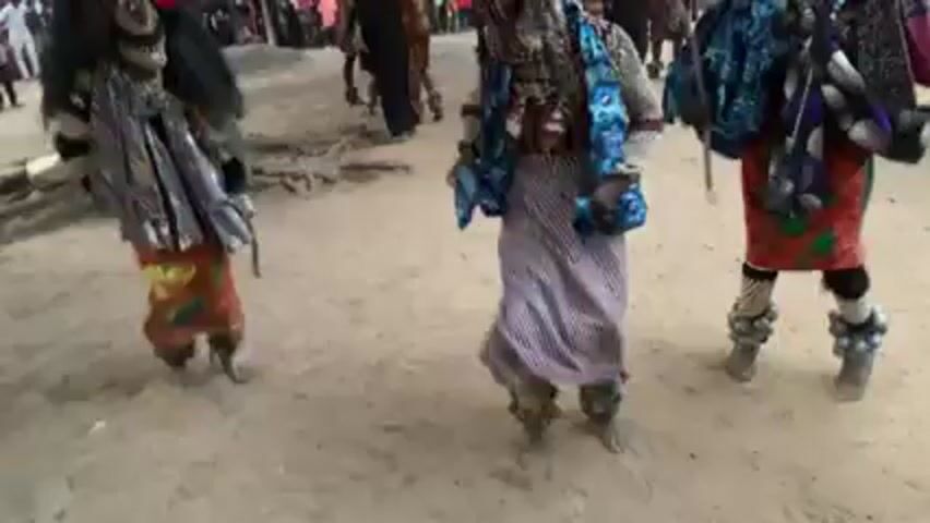 The video shows a group of traditionally dressed people performing in what appears to be a public space, likely a village. Three primary figures are heavily costumed in masks and colorful, patterned clothing, including skirts and tops. They are moving energetically, suggesting a dance or ritualistic performance. One of the figures stumbles or falls, causing the other two to move around them. Onlookers, including adults and children, are visible in the background, watching the performance. The ground is sandy and there are some trees in the vicinity. The overall atmosphere seems festive and celebratory.